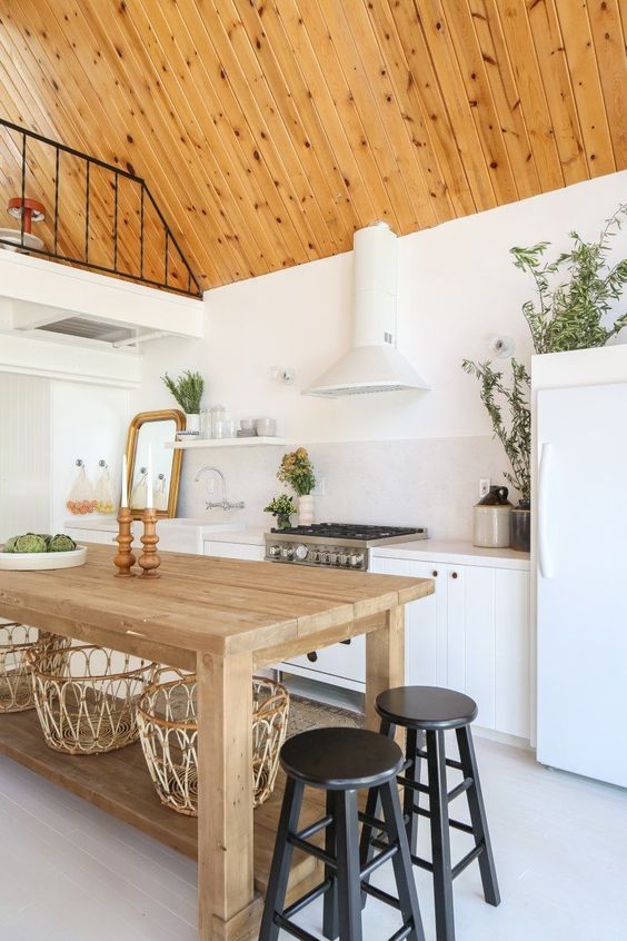 a pretty modern barn kitchen with a wood clad ceiling, white planked cabinets, a stained table, black stools and lots of greenery