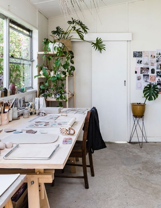 a simple and pretty art studio with a large table, a chair, potted plants, lots of art and art supplies is amazing