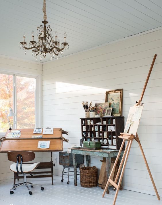 a small art nook with an easel, a chair, a shabby chic table with a shelving unit, an easel, a crystal chandelier