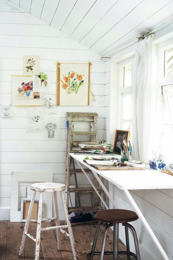 a small rustic art nook by the window with a wall-mounted table, stools, a ladder, some art and art supplies