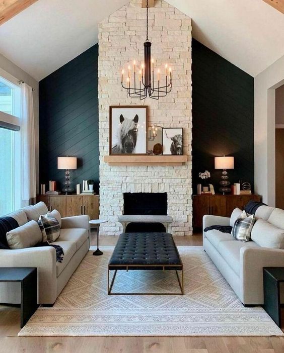 a sophisticated modern barn living room with a fireplace clad with faux stone, neutral seating furniture, a printed rug, a black ottoman, table lamps and a chic chandelier