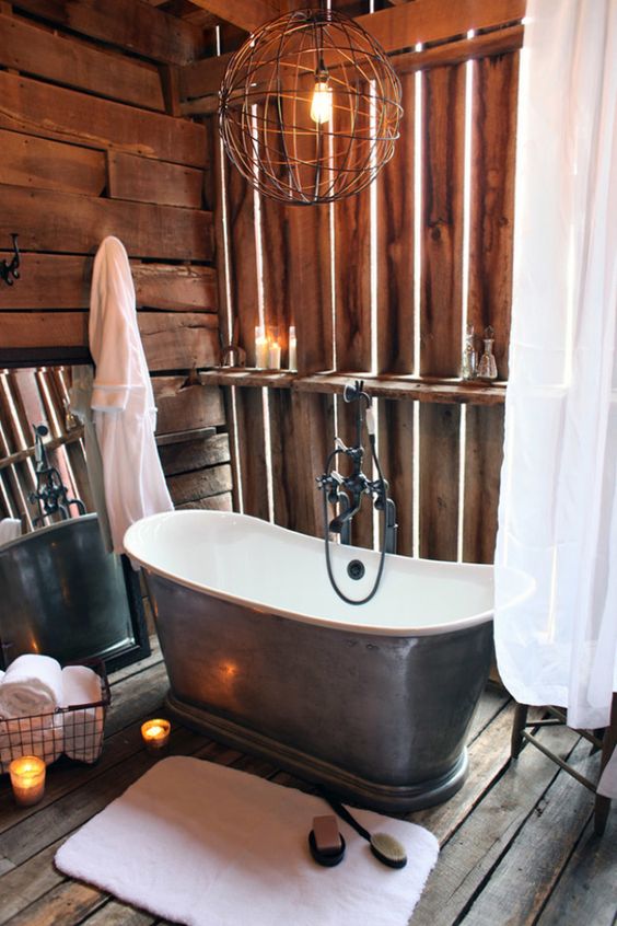 a vintage barn bathroom with wood all over it, a vintage metal tub, a sphere chandelier, candles and a wire basket