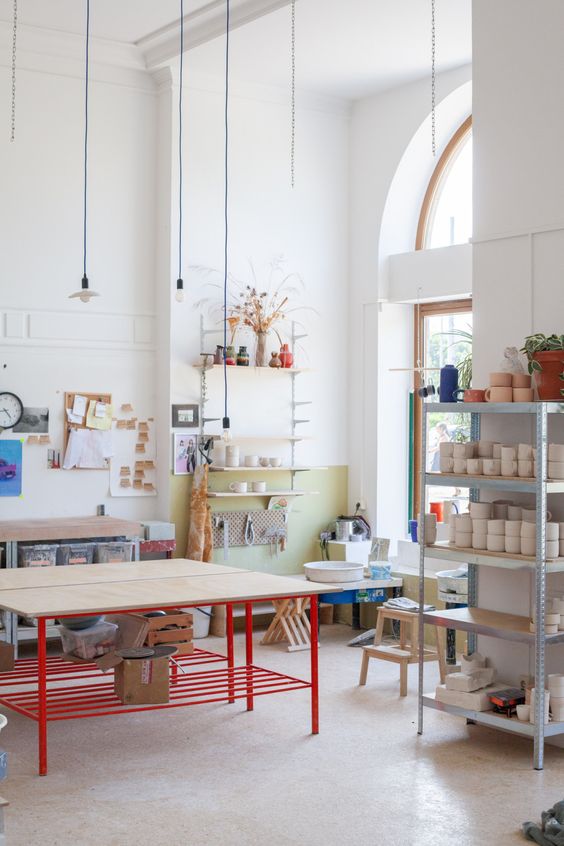 an airy Scandinavian home artist studio with large tables, an open storag eunit, some benches, stools and shelves, touches of color and greenery
