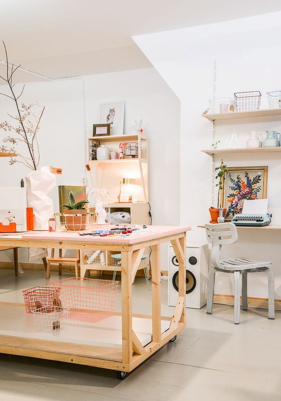an airy home artist and craft studio with open shelves and decor, chairs and stools, a large table, some artwork and some plants