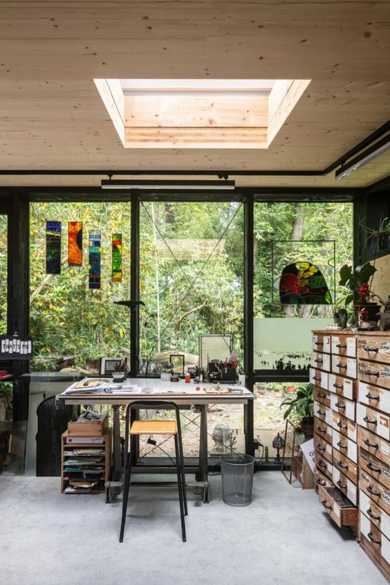 an art studio with a glazed wall, a file cabinet, a metal desk and a stool, some storage units, colorful artwork and potted plants