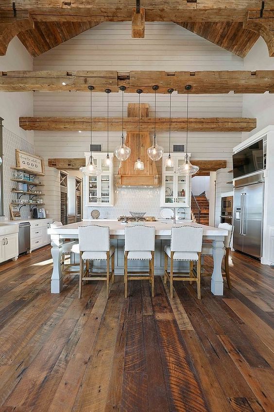 an elegant white barn kitchen with planked walls, stained wooden beams, shaker style cabinets, a vintage kitchen island and white stools