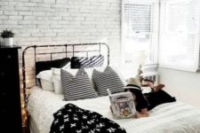 a Scandinavian bedroom with a faux white brick wall, a black metal bed and dresser, lights and monochromatic bedding