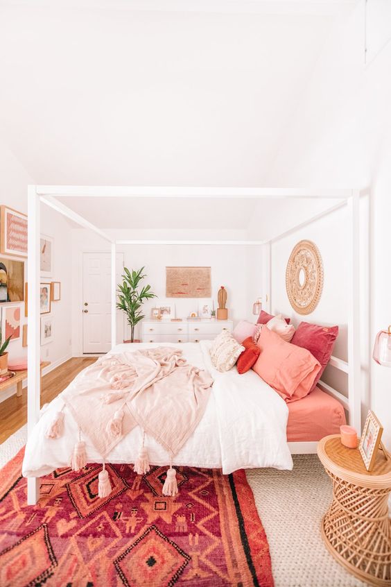 a beautiful white boho bedroom with a canopy bed, a rattan table and a gallery wall, bright bedding and a red rug is cool