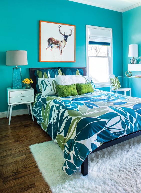 a bright turquoise bedroom with a dark bed and bright bedding, a white nightstand with a lamp, a white chair and pillows and an artwork