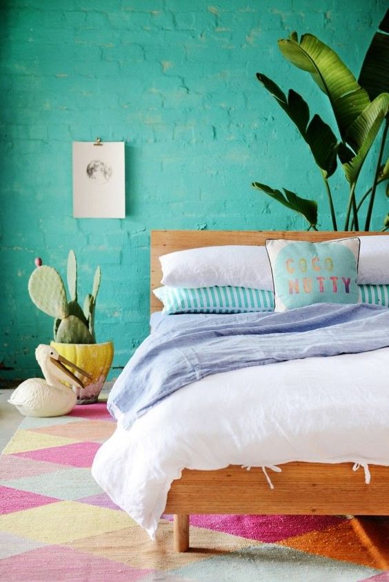a bright tropical bedroom with a turquoise brick wall, a stained bed with bright bedding, a colorful rug and potted plants