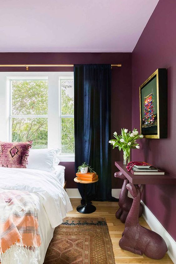 a catchy purple bedroom with bold walls, a bed, a console table on animal like legs, navy curtains and a bold artwork