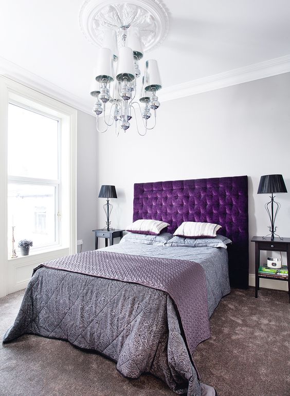 a chic bedroom with a bright purple bed, dark nightstands and lamps, a catchy vintage chandelier and grey and purple bedding