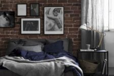 a chic moody bedroom with a faux brick wall, a cork bench, a dark bed and a monochromatic gallery wall