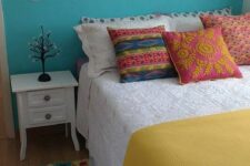 a colorful bedroom with a turquoise accent wall, a bed with colorful bedding, a shelf with decor, a white nightstand and a crochet rug