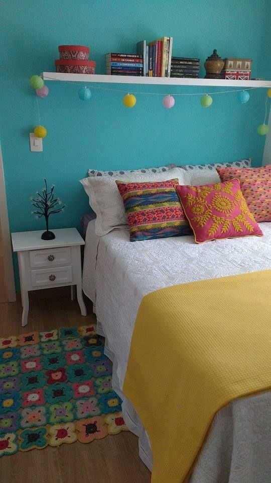 a colorful bedroom with a turquoise accent wall, a bed with colorful bedding, a shelf with decor, a white nightstand and a crochet rug