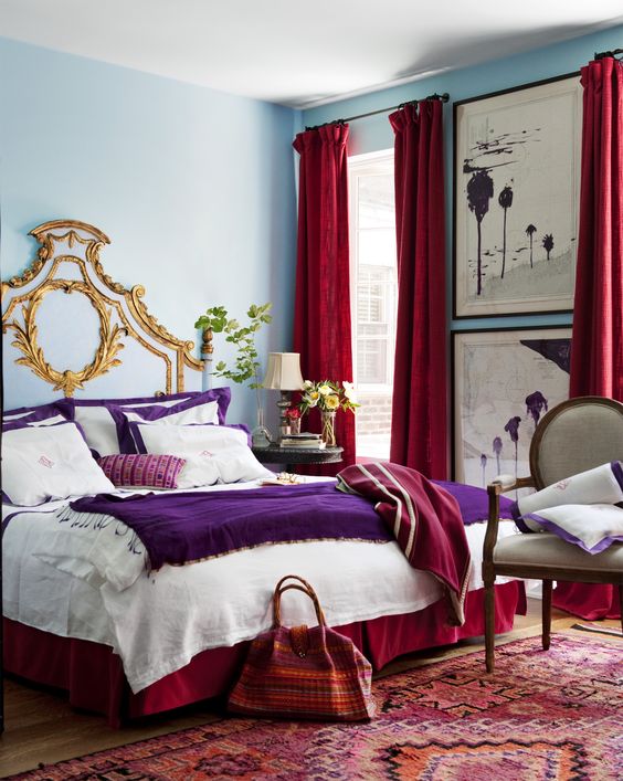 a colorful bedroom with blue walls, a bed with purple and white bedding, a vintage chair, red curtains, a red printed rug and a red blanket