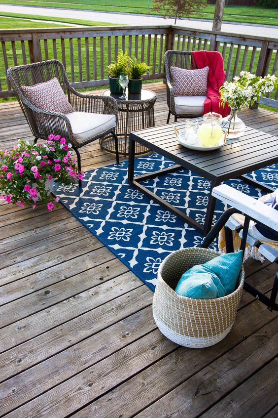 a colorful eclectic deck with a bright rug, wicker and wooden furniture, colorful textiles and bright blooms in planters
