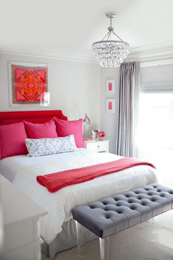 a glam neutral bedroom with a red upholstered bed, neutral and red bedding, a grey upholstered bench and a crystal chandelier