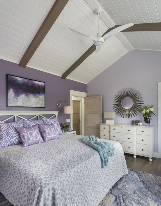a lavender and white bedroom with a purple statement wall, white furniture, a bold artwork, a sunburst mirror and some lavender bedding