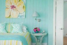 a lovely cottage bedroom with a turquoise accent wall, a bed with turquoise and yellow bedding, a bright artwork, a mint nightstand and a sconce