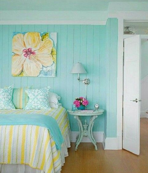a lovely cottage bedroom with a turquoise accent wall, a bed with turquoise and yellow bedding, a bright artwork, a mint nightstand and a sconce