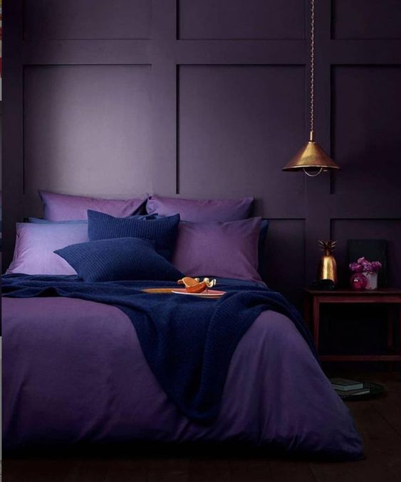 a moody monochromatic bedroom with deep purple paneled walls, purple and navy bedding, a wooden nightstand and a metal pendant lamp