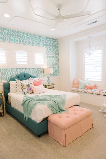 a pretty mid-century modern bedroom with a turquoise accent wall, a teal bed with colorful bedding, a peachy pink ottoman and a windowsill bench