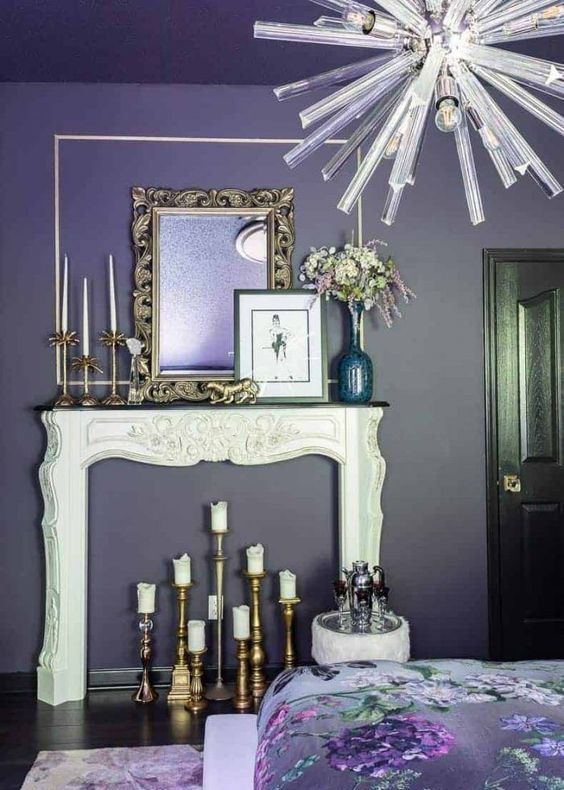 a purple and white bedroom with a faux fireplace, some gold candleholders and a mirror ina frame, a chandelier with bulbs and floral bedding