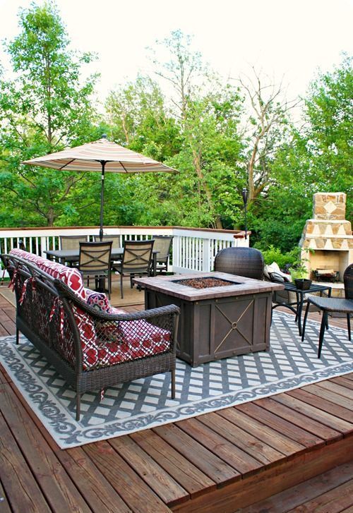 a rustic deck with dark wicker furniture, an umbrella with a dining zone, a fire pit in the center and a fireplace