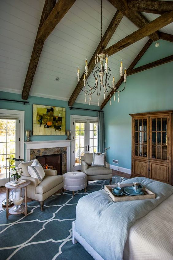 a vintage attic bedroom with wooden beams, turquoise walls, a bed with light blue bedding, a stained bookcase, a fireplace and some seating furntiure