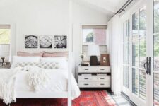 a white boho bedroom with an attic ceiling, a white canopy bed with neutral bedding, a bold red rug and some nightstands