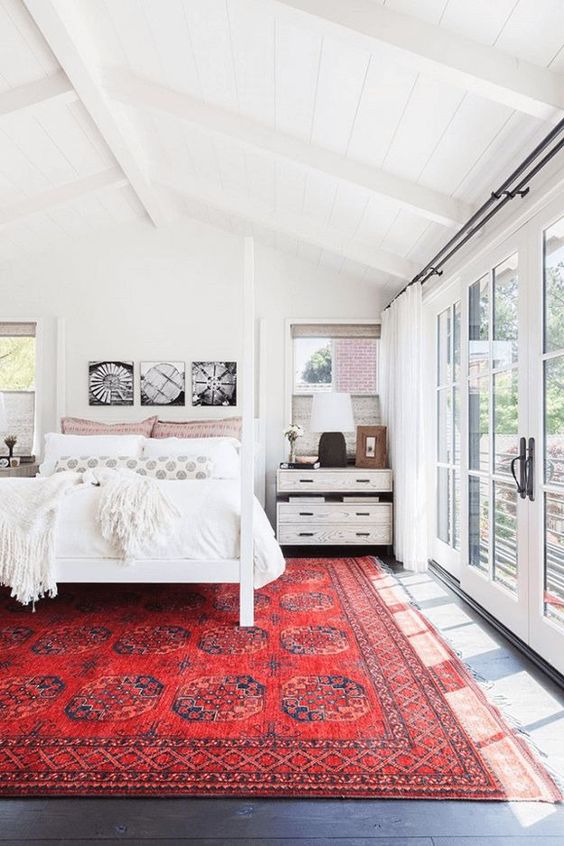 a white boho bedroom with an attic ceiling, a white canopy bed with neutral bedding, a bold red rug and some nightstands