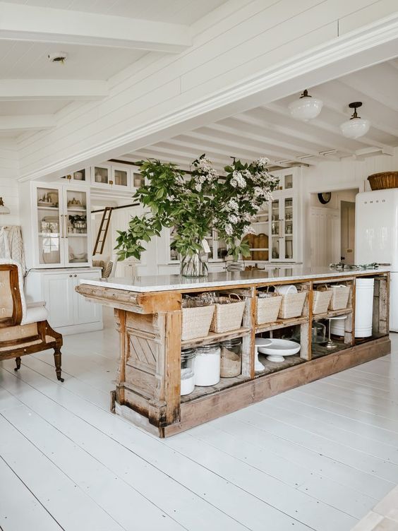 a white cottage kitchen with glass cabinets, a large antique kitchen island with plenty of storage space and greenery