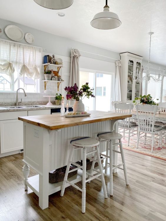 a white farmhouse kitchen filled with light, with a small wooden kitchen island with a stained countertop and tall white stools