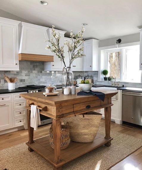a white farmhouse kitchen with shaker style cabinets, black countertops, a hood with a wooden trim and a stained kitchen island on refined legs