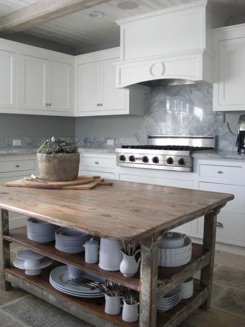 a white kitchen with a marble tile backsplash and countertops, a vintage hood and a stained wooden kitchen island that is a tabke