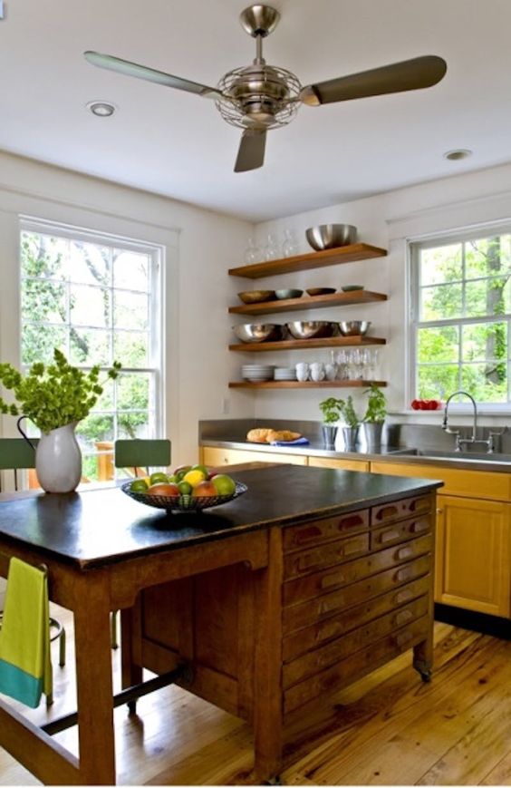 a yellow kitchen with dark countertops, open shelves instead of upper cabinets, a vintage desk as a kitchen island and green stools