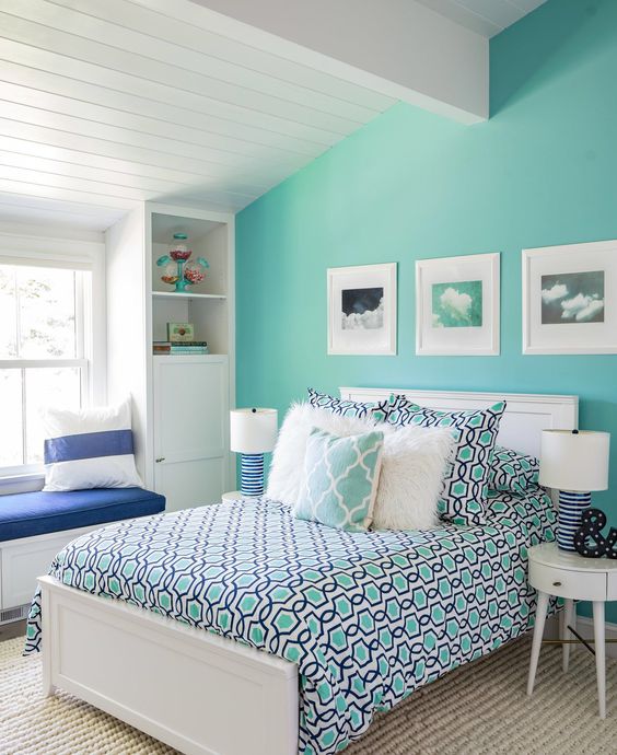 an attic bedroom with a turquoise accent wall, a white bed and colorful bedding, a windowsill bench and built in storage units