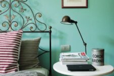 an eclectic bedroom with a turquoise accent wall, a forged bed and a round nightstand, a black table lamp and some magazines