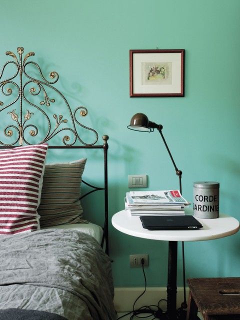 an eclectic bedroom with a turquoise accent wall, a forged bed and a round nightstand, a black table lamp and some magazines