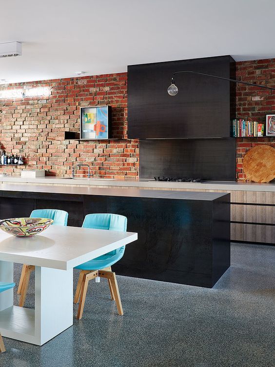an eclectic kitchen with sleek light and dark cabinets, a red brick wall and bright turquoise chairs