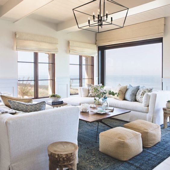 a beach living room in navy, blues and tan, with upholstered furniture, leather ottomans, tan Roman shades and printed pillows