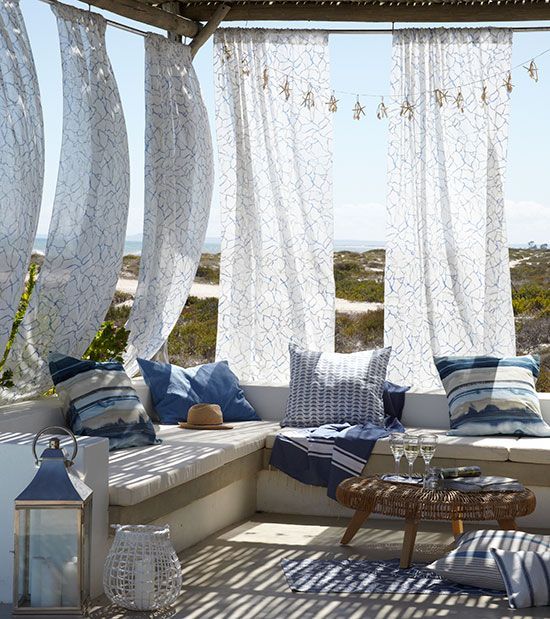 a beach patio with printed curtains, a bench with bright blue pillows, a wicker table and a candle lantern is very welcoming