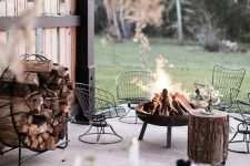 a beautiful rustic outdoor space with a simple wrought iron fire bowl on legs, metal chairs, a tree sutmp and a firewood stand