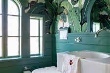 a bold retro bathroom with banana leaf wallpaper on the ceiling, emerald paneling, a floating vanity and arched windows