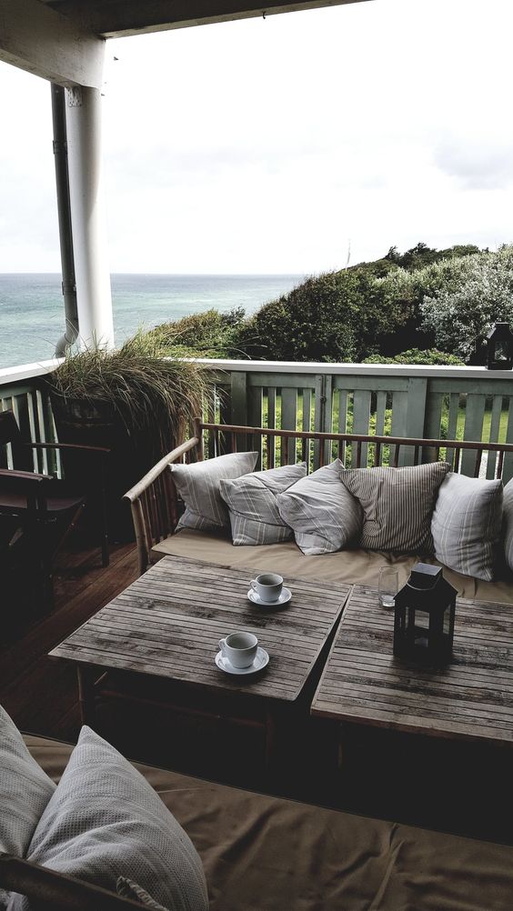 a casual seaside patio with simple wooden furniture, neutral printed pillows, candle lanterns and potted grasses