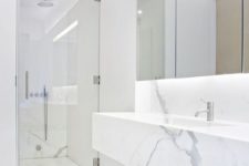 a clean minimalist space done with whte marble, a mirror with lights, a shower space and a marble vanity