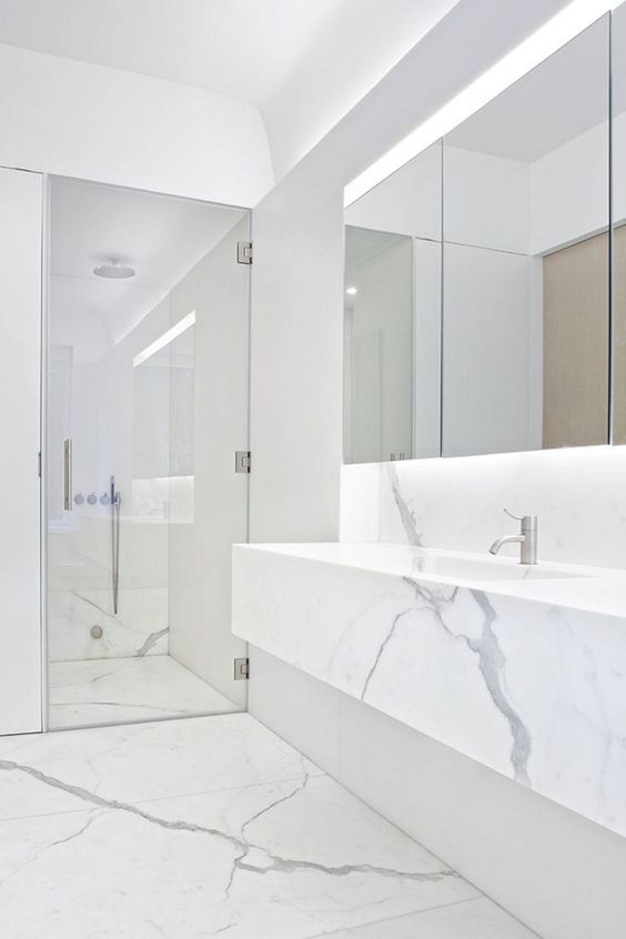 a clean minimalist space done with whte marble, a mirror with lights, a shower space and a marble vanity