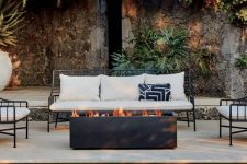 a cool and welcoming outdoor space with white forged furniture, a black fire bowl of a rectangular shape next to the pool