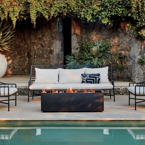 a cool and welcoming outdoor space with white forged furniture, a black fire bowl of a rectangular shape next to the pool